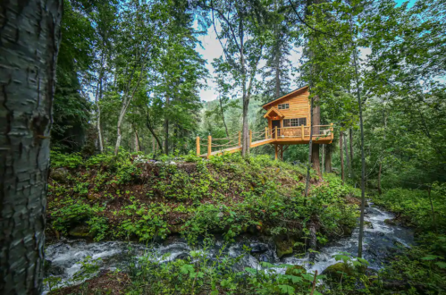 A wooden treehouse perched on a hillside, surrounded by lush greenery and a flowing stream below.