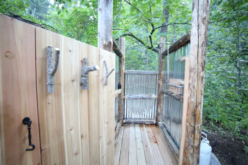 A rustic outdoor shower area with wooden walls and a metal door, surrounded by lush greenery.