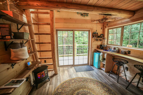 Cozy wooden interior of a cabin with a ladder, kitchenette, and large windows overlooking a forested area.