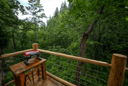 A wooden deck overlooking a lush green forest, featuring a small table with a grill and cooking utensils.