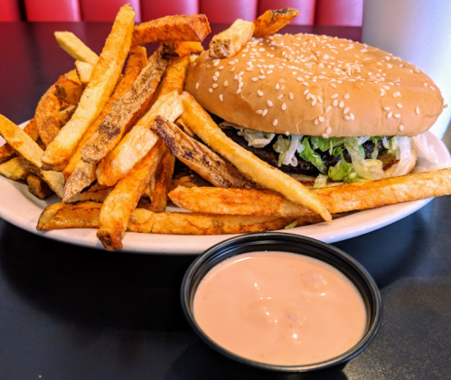 A plate of crispy French fries next to a sesame seed burger topped with lettuce, served with a small cup of sauce.