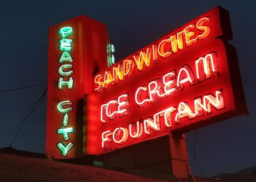 Neon sign for "Peach City" featuring "Sandwiches," "Ice Cream," and "Fountain" illuminated at night.