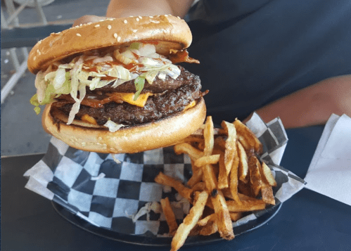 A hand holding a large cheeseburger with lettuce, bacon, and a side of crispy fries on a checkered plate.