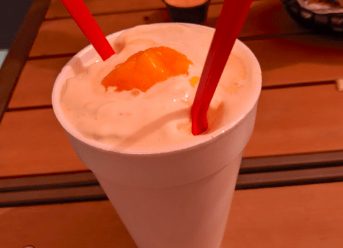 A large foam cup filled with a creamy drink topped with whipped cream and a slice of orange, with two red straws.