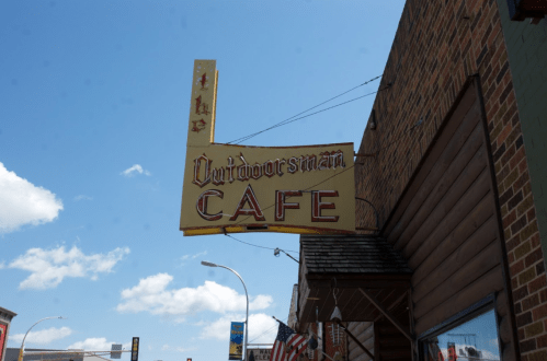 Sign for "The Outdoorsman Cafe" against a blue sky with clouds, featuring a vintage design and bright colors.