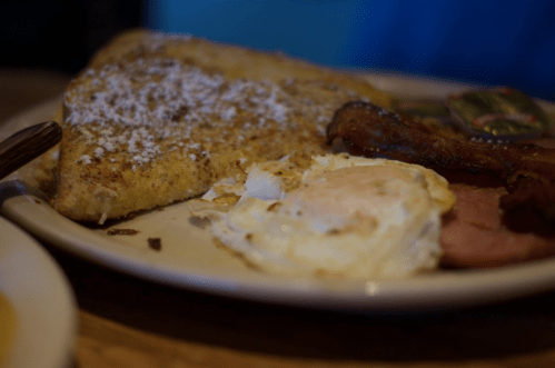 A plate of breakfast featuring French toast, fried eggs, bacon, and sausage, dusted with powdered sugar.