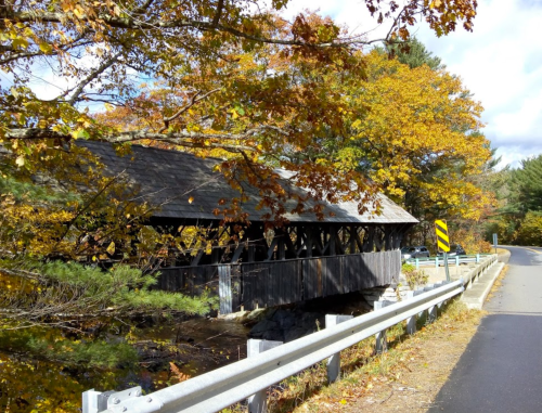 A covered bridge surrounded by autumn trees, with a road and guardrail in the foreground.