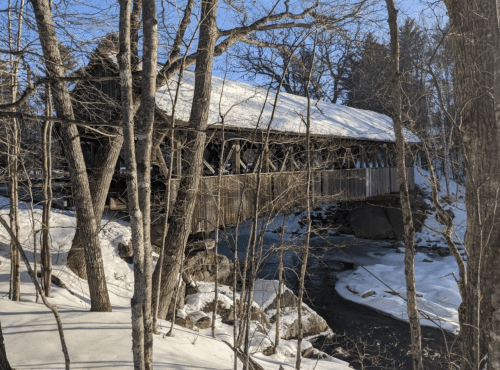 A snow-covered wooden bridge spans a stream, surrounded by bare trees in a winter landscape.