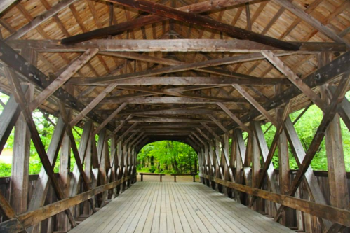 Interior view of a wooden covered bridge with a sloped roof and green trees visible in the background.