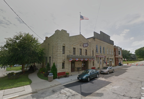 Historic stone building with an American flag, featuring decorative bunting and parked cars in a small town setting.
