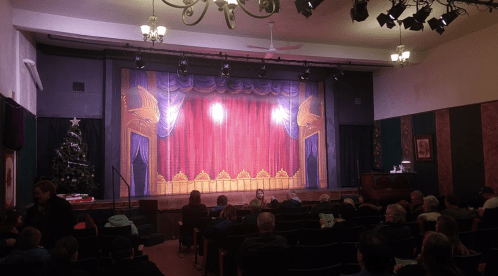 A theater stage with a red curtain and decorative backdrop, filled with audience members waiting for a performance.