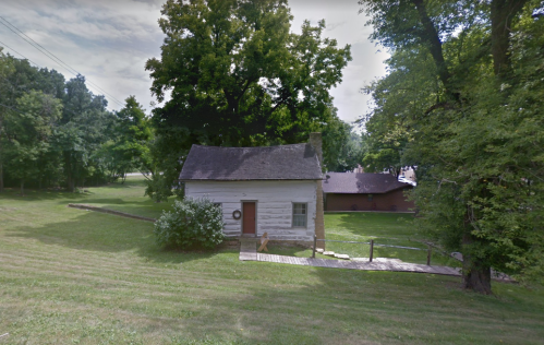 A small, white house surrounded by trees and grass, with a wooden walkway leading to it.