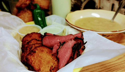 A plate featuring crispy fried patties, slices of smoked meat, and two dipping sauces, with a bowl of soup in the background.