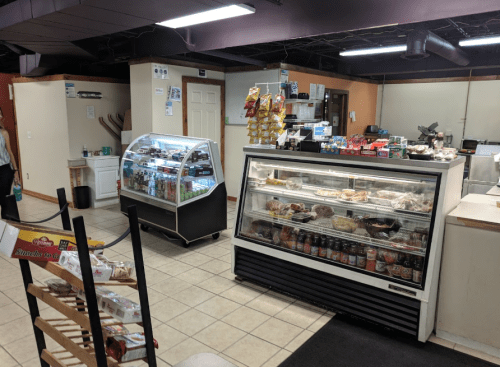 A small deli interior featuring a glass display case with food, drinks, and snacks, with a checkout area in the background.