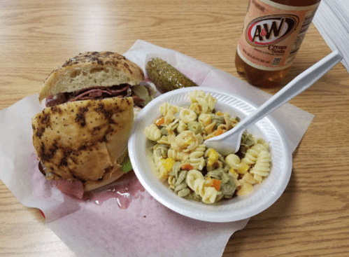 A plate with a sandwich, pasta salad, a pickle, and a bottle of A&W root beer on a wooden table.