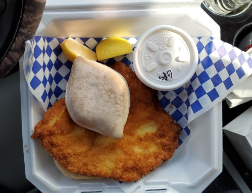 A breaded fried fish fillet with a lemon wedge and a side of creamy sauce in a takeout container.