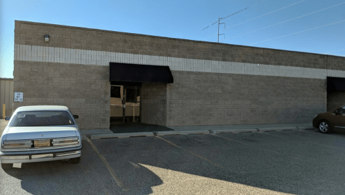 A plain, beige building with a black awning and two parked cars in front, under a clear blue sky.