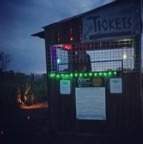 A dimly lit ticket booth with colorful lights, set against a twilight sky, promoting a haunted attraction.