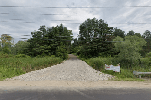 Gravel road leading into a wooded area, surrounded by greenery and power lines overhead.
