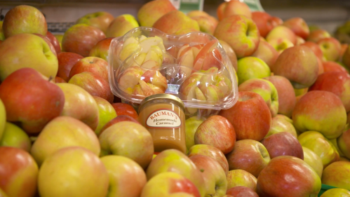 A clear container of sliced apples sits atop a pile of red and green apples, next to a jar of homemade caramel.