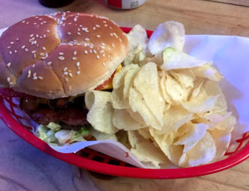 A sesame seed bun burger with lettuce, tomato, and bacon, served with a side of potato chips in a red basket.