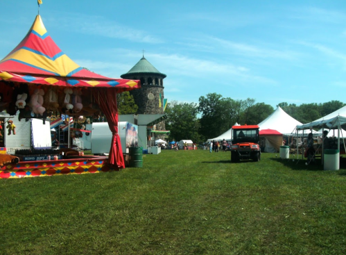 A fairground scene with colorful tents, a tower in the background, and a grassy path lined with vendor stalls.