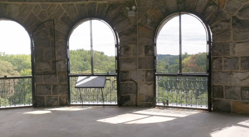 Interior of a stone tower with three arched windows, showcasing a view of trees outside. Natural light filters in.