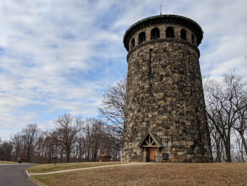 A tall, round stone tower with a green roof, surrounded by bare trees and a cloudy sky.