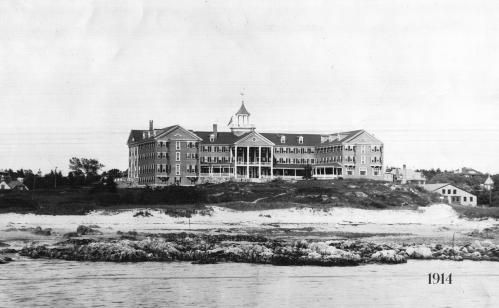 Historic black-and-white photo of a large hotel by the shore, dated 1914, with rocky beach in the foreground.