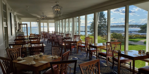Bright dining area with wooden tables and chairs, large windows overlooking a scenic waterfront view.