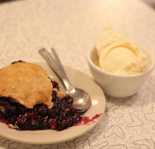 A slice of berry pie with a scoop of vanilla ice cream on a plate, accompanied by a spoon.
