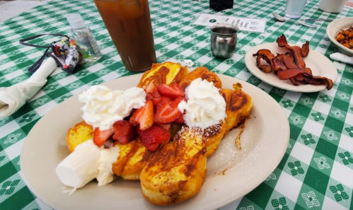 A plate of French toast topped with strawberries and whipped cream, alongside a side of bacon and a drink.