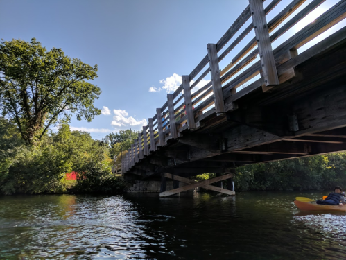 A wooden bridge spans over a calm river, surrounded by lush greenery and a clear blue sky. A kayaker is visible below.