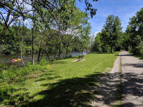 A peaceful riverside scene with a grassy area, a bench, and a path alongside the water, surrounded by trees.