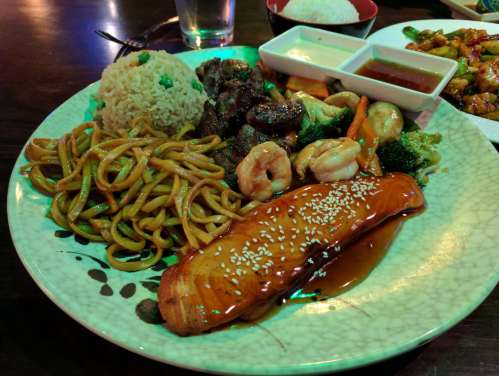 A colorful plate featuring rice, noodles, shrimp, grilled chicken, vegetables, and sesame-glazed salmon.