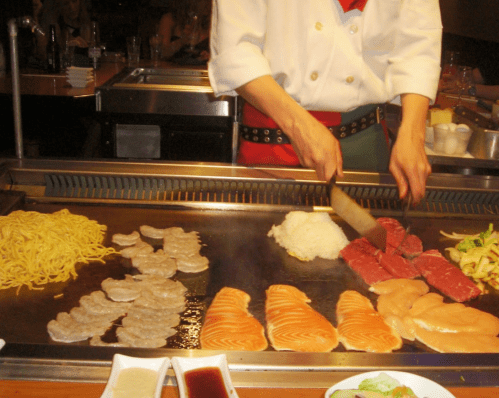 A chef prepares a variety of grilled foods, including shrimp, salmon, chicken, and vegetables on a teppanyaki grill.