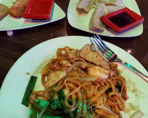 A plate of stir-fried noodles with chicken and vegetables, accompanied by dumplings and dipping sauces on the side.