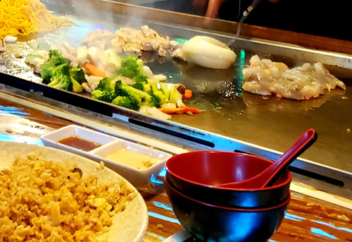 A sizzling teppanyaki grill with vegetables, seafood, and chicken, alongside a bowl of fried rice and dipping sauces.