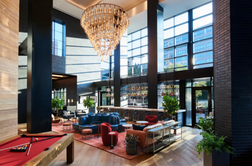 Modern lobby with a chandelier, stylish seating, and large windows, featuring a bar and greenery.