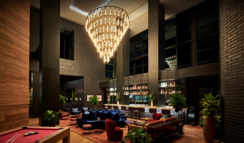 A stylish lounge with a large chandelier, modern seating, and a bar lined with bottles, surrounded by greenery.