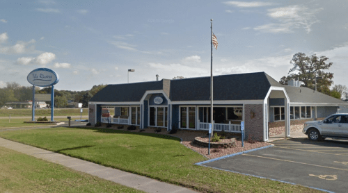 A brick and siding restaurant building with a sign, flag, and grassy area, set against a clear blue sky.