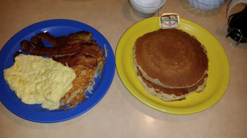 A blue plate with scrambled eggs, bacon, and hash browns next to a yellow plate with stacked pancakes and syrup.