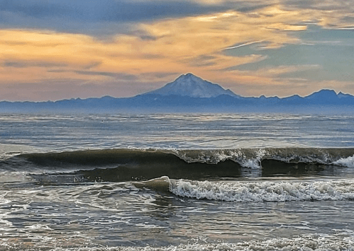 A serene beach scene with gentle waves and a distant snow-capped mountain under a colorful sunset sky.