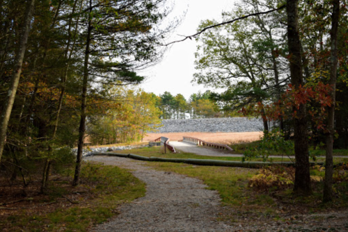 A gravel path leads through trees to a dam, with a grassy area and stone structure in the background.