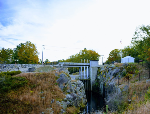 A dam structure surrounded by rocky terrain and trees, with a small building and an American flag in the background.