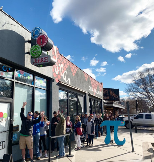 A long line of people waiting outside a colorful shop on a sunny day, with a blue pi symbol in the foreground.