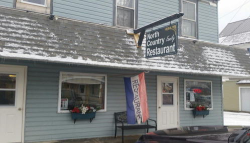Exterior of North Country Restaurant with a sign, open flag, and snow on the roof. Flowers decorate the windows.