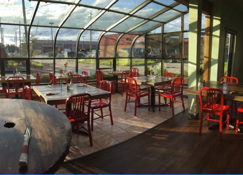 Bright, airy restaurant interior with red chairs, wooden tables, and large windows showcasing a sunny outdoor view.