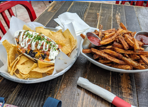 A plate of nachos with toppings and a side of crispy fries served with dipping sauce on a wooden table.