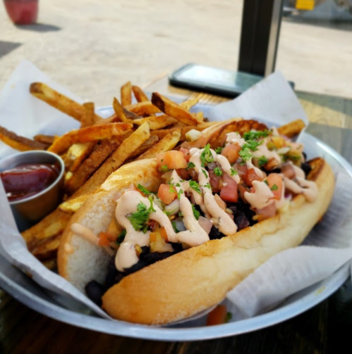 A hot dog topped with diced tomatoes and sauce, served with crispy fries and a small cup of ketchup.